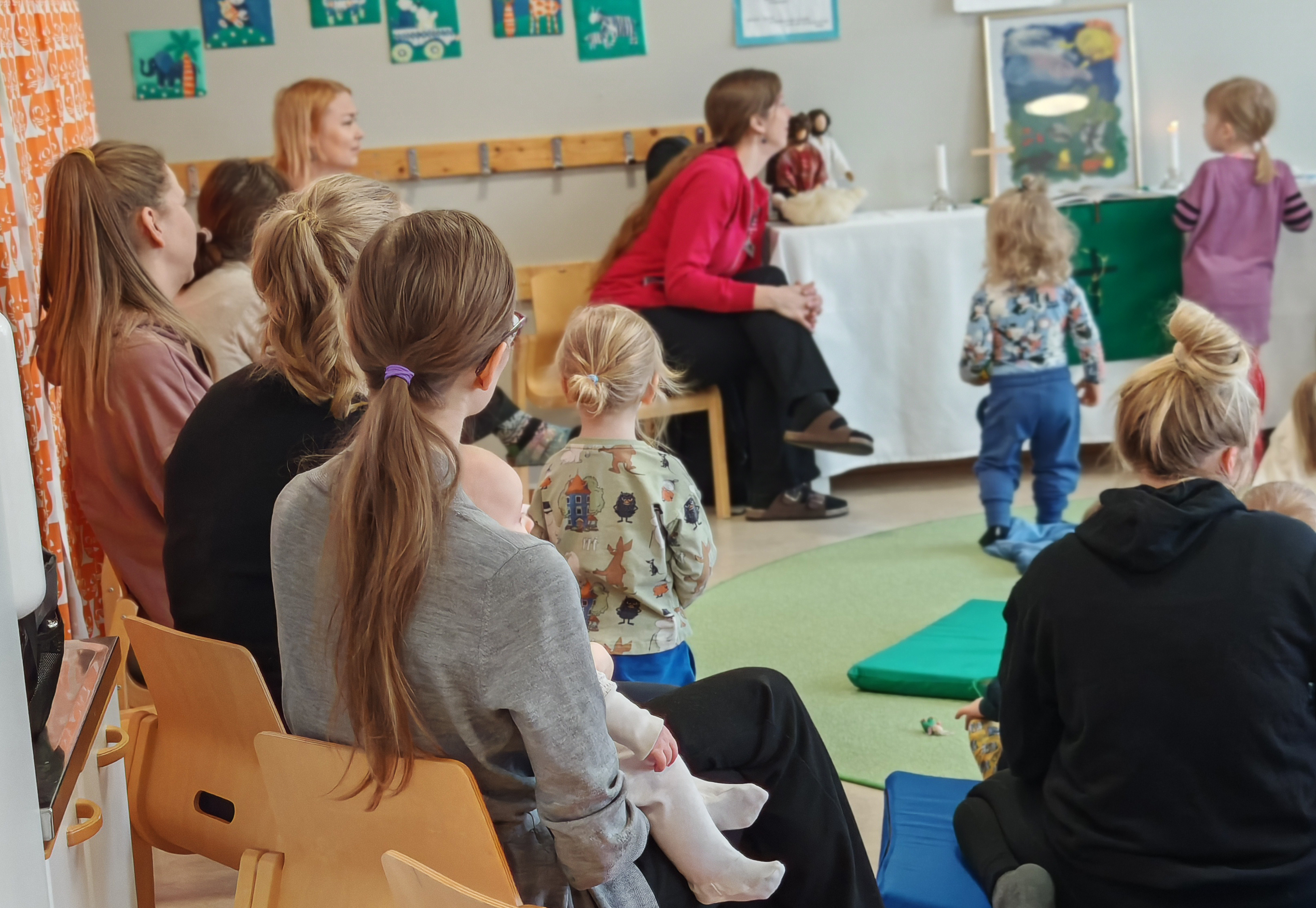Valokuva perhekerhon hartaushetkestä. Edustalla selkien takaa kuvattuja kerhokävijöitä istumassa tuoleilla ...