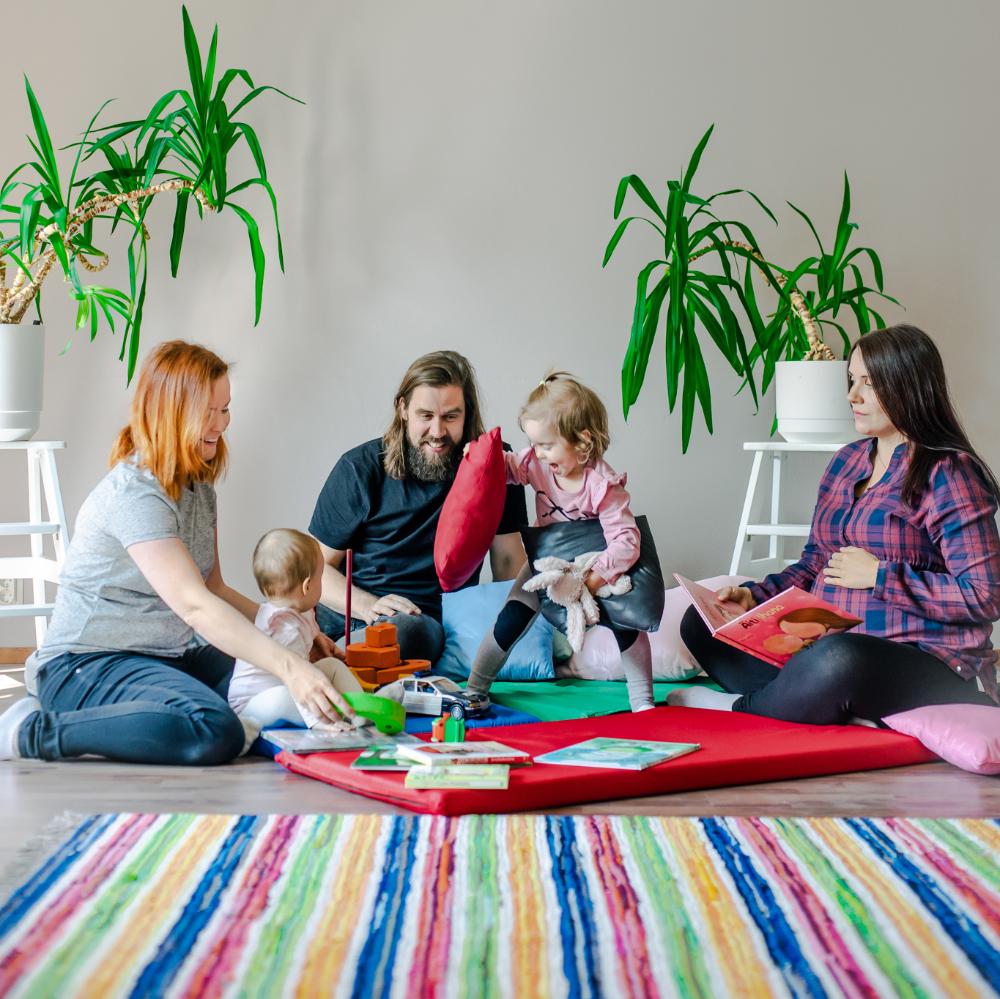 Kaksi lasta ja kolme aikuista leikkivät lattialla värikkäiden mattojen ja tyynyjen päällä.