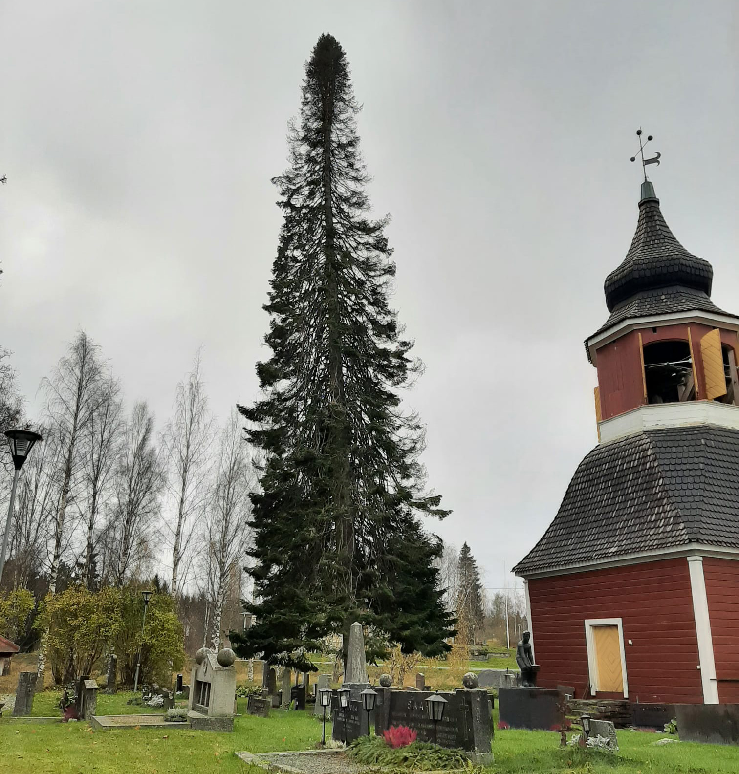 Pilvisellä kesäsäällä otettu valokuva Kuhmalahden hautausmaalla sijaitsevasta kellotapulista ja sen vieress...