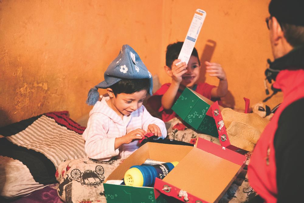 Kaksi lasta avaamassa Joulun Lapsi paketteja. Iloiset ilmeet ja värikäs kuva.